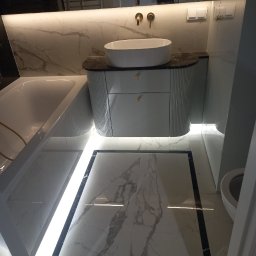 USŁUGI REMONTOWO-BUDOWLANE VOLODYMYR NAVROTSKYY - Elegancka łazienka z wanną, umywalką na szafce z ryflowanymi frontami i marmurową podłogą z czarną ramą, oświetlona ledami. Widoczne odbicie osoby w lustrze.