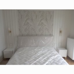 USŁUGI REMONTOWO-BUDOWLANE VOLODYMYR NAVROTSKYY - Elegancka sypialnia z łóżkiem zabezpieczonym folią, pasująca tapeta w liście, symetryczne szafki nocne i lampki ścienne. Grzejnik żeberkowy w tle.