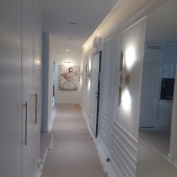 USŁUGI REMONTOWO-BUDOWLANE VOLODYMYR NAVROTSKYY - Biały korytarz z szafami w zabudowie, sztukaterią i złotymi lampami ściennymi. Na końcu korytarza widoczny obraz z baletnicą.