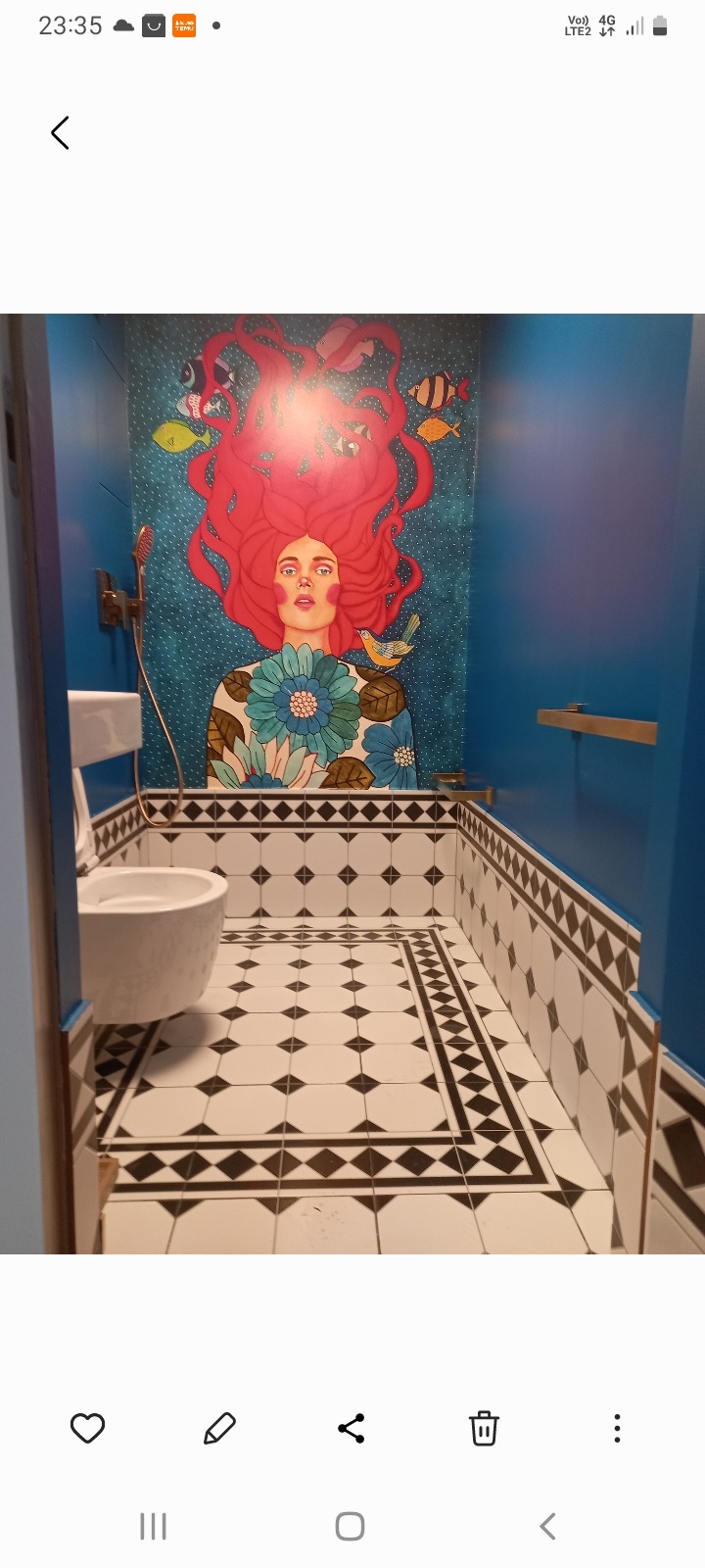 Niebieska łazienka z artystycznym muralem: kobieta z czerwonymi włosami i rybami. Podłoga w biało-czarną szachownicę. Toaleta widoczna z boku.