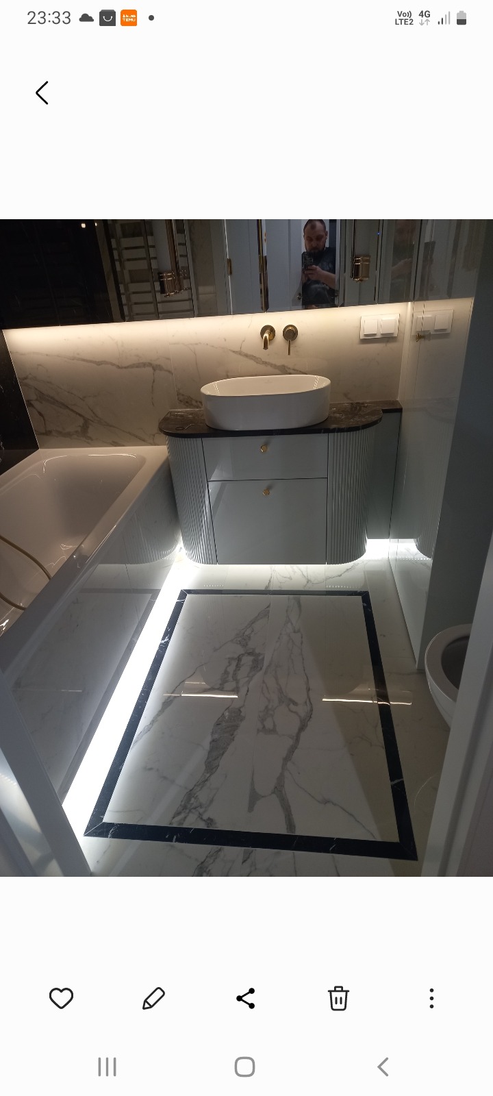 Elegancka łazienka z wanną, umywalką na szafce z ryflowanymi frontami i marmurową podłogą z czarną ramą, oświetlona ledami. Widoczne odbicie osoby w lustrze.