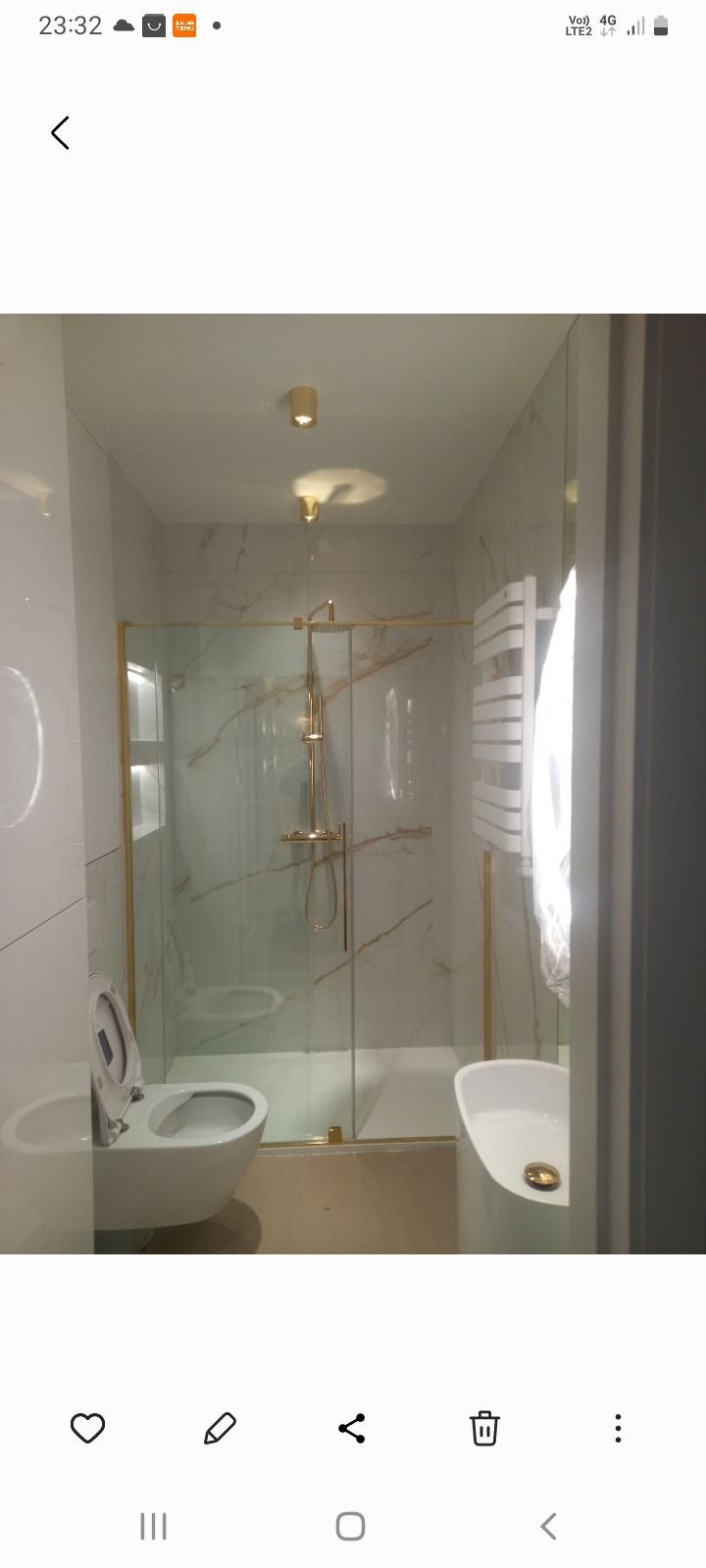 Nowoczesna łazienka z kabiną prysznicową w złotej ramie, marmurowymi płytkami i białymi sanitariatami. Widoczne oświetlenie punktowe i grzejnik.