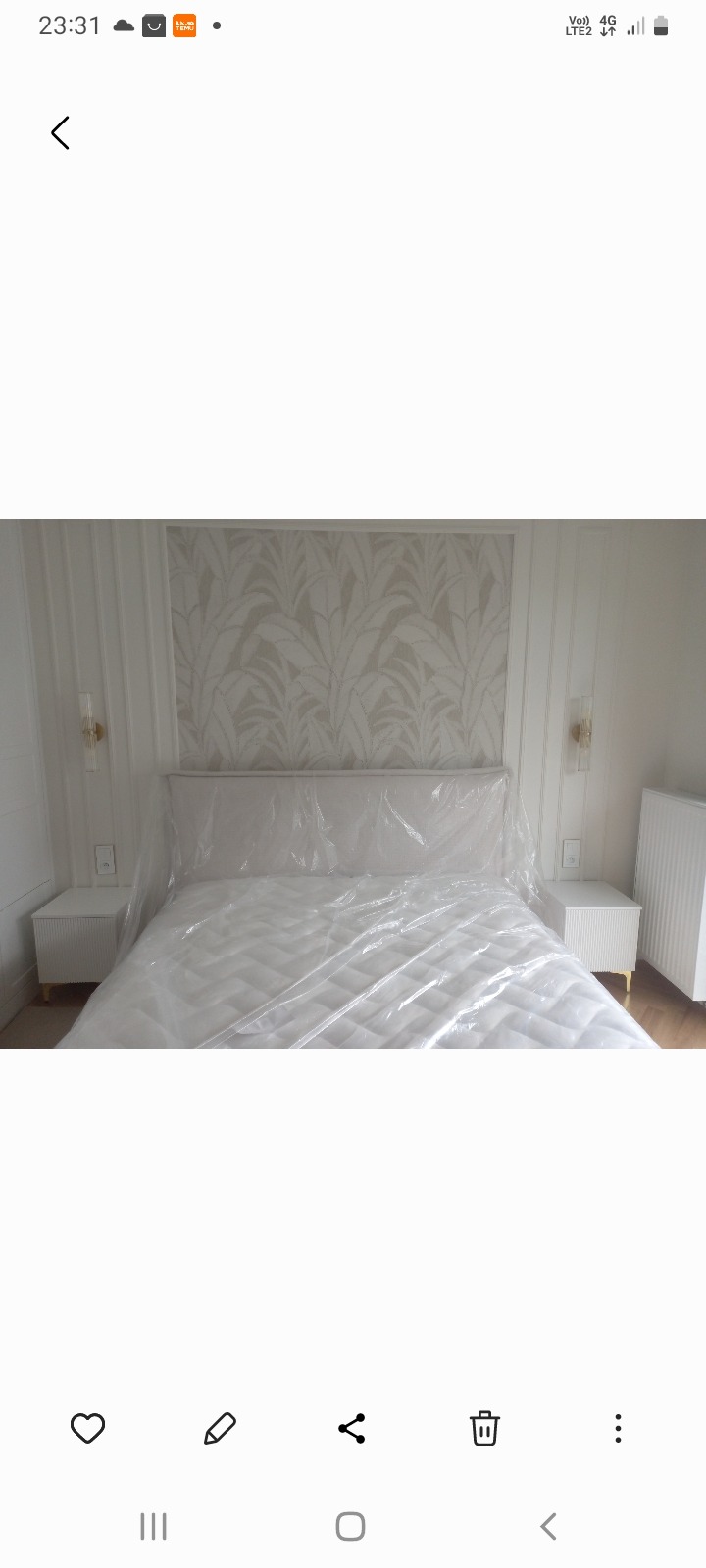 Elegancka sypialnia z łóżkiem zabezpieczonym folią, pasująca tapeta w liście, symetryczne szafki nocne i lampki ścienne. Grzejnik żeberkowy w tle.