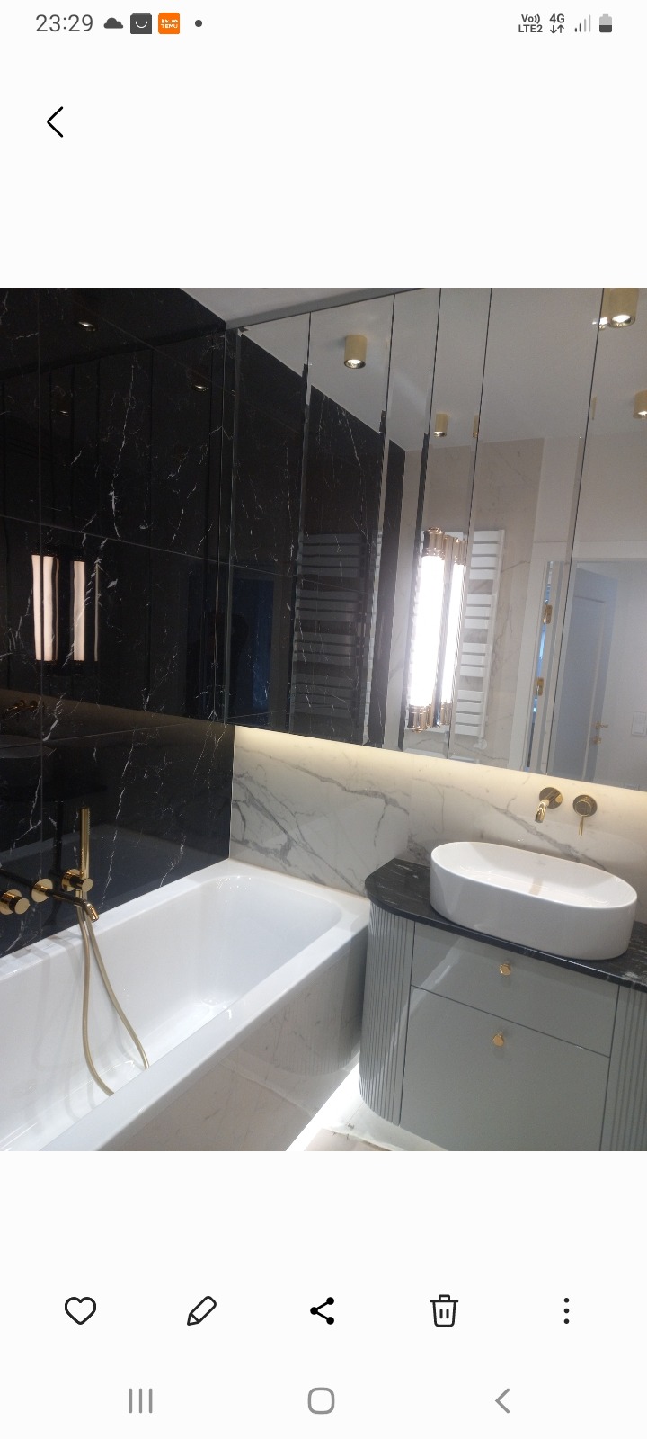 Elegancka łazienka z wanną, umywalką nablatową i czarnymi płytkami imitującymi marmur. Lustro z pionowymi pasami. Złote detale armatury i oświetlenia.