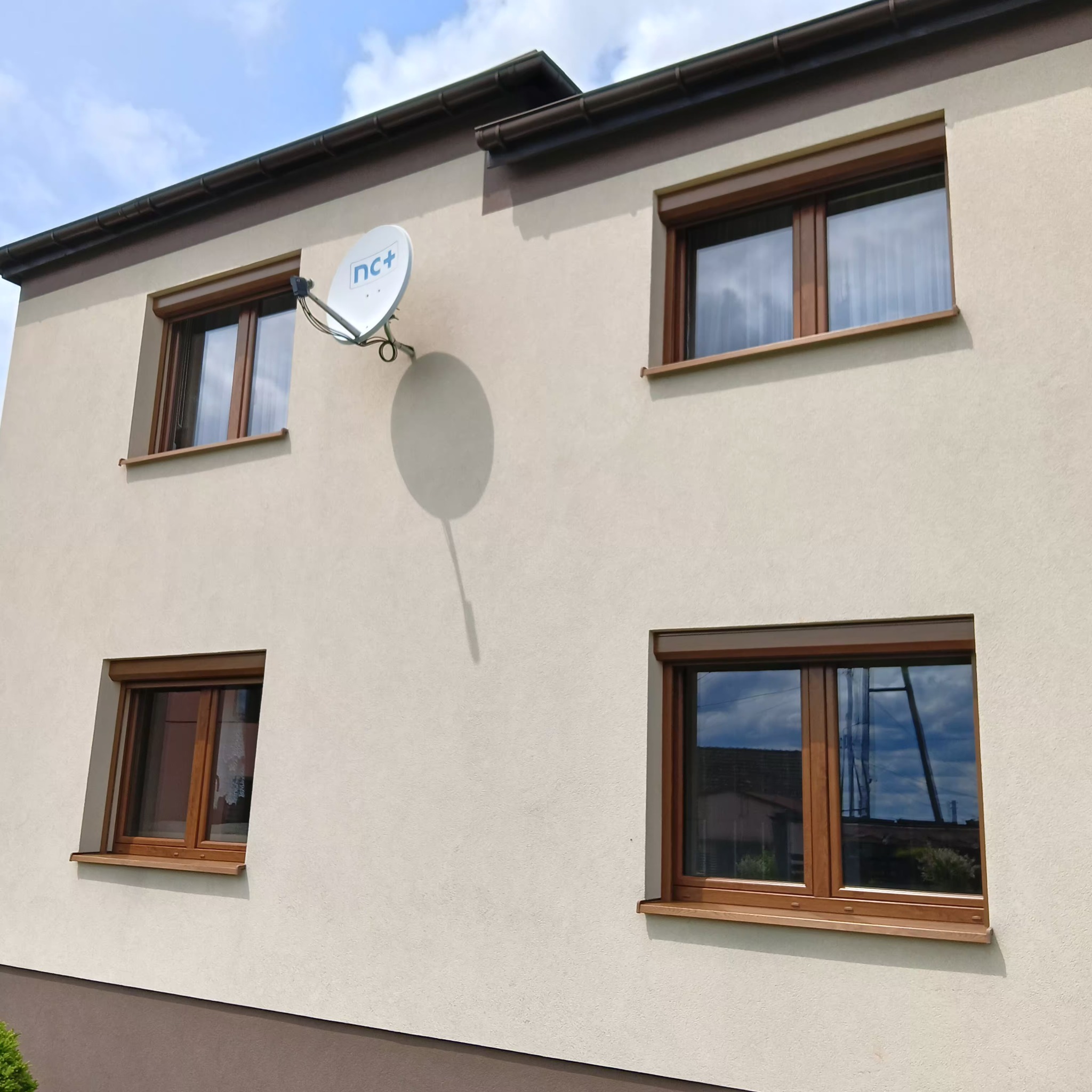 Elewacja domu z oknami z brązowymi roletami zewnętrznymi, częściowo opuszczonymi. Na ścianie widoczna antena satelitarna.