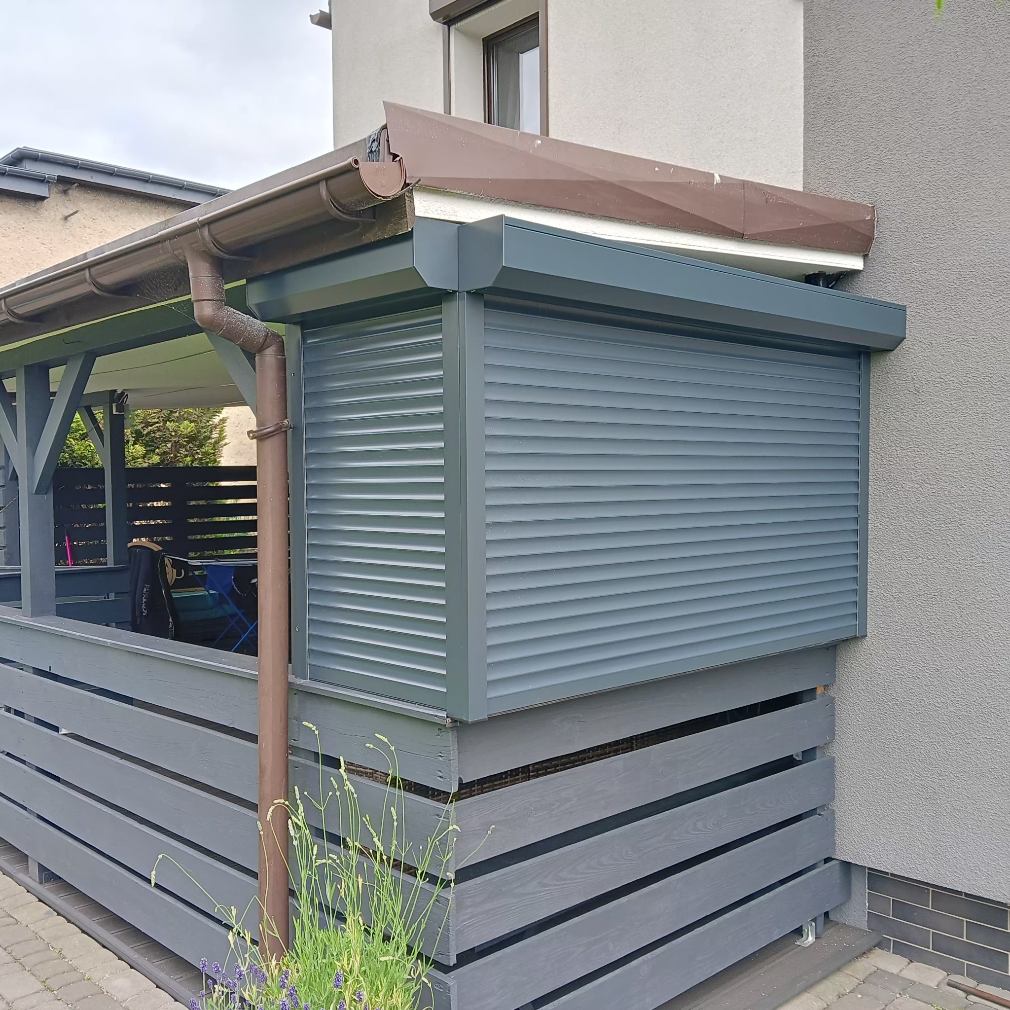 Szara roleta zewnętrzna zamontowana na drewnianej konstrukcji tarasu, z widocznym fragmentem rynny i elewacji budynku w tle. Na pierwszym planie rośliny.