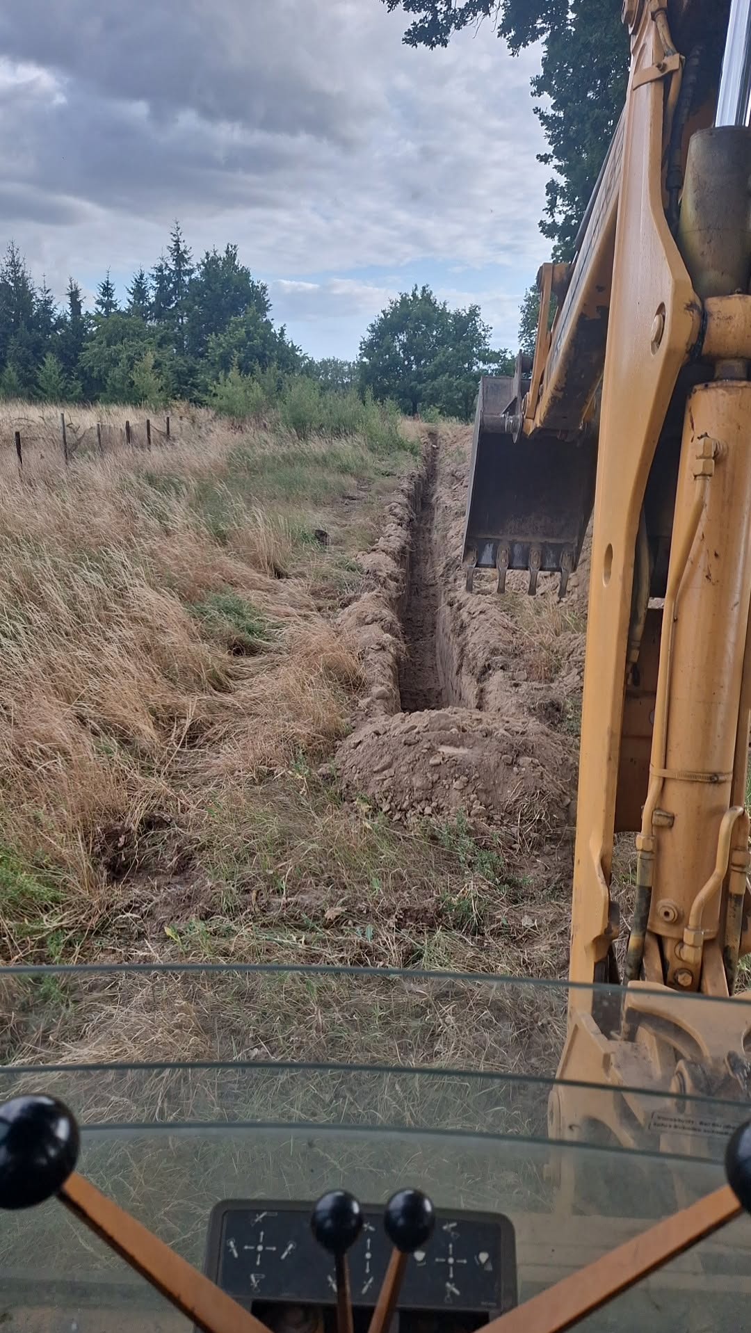 Wąski wykop w ziemi, widoczny z kabiny koparki z żółtym ramieniem. W tle trawa i drzewa pod pochmurnym niebem.