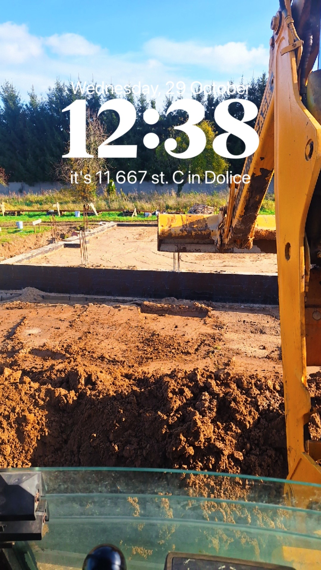 Żółta koparka przy wykopie fundamentów z wystającymi prętami zbrojeniowymi, widok z kabiny operatora, w tle drzewa i błękitne niebo.