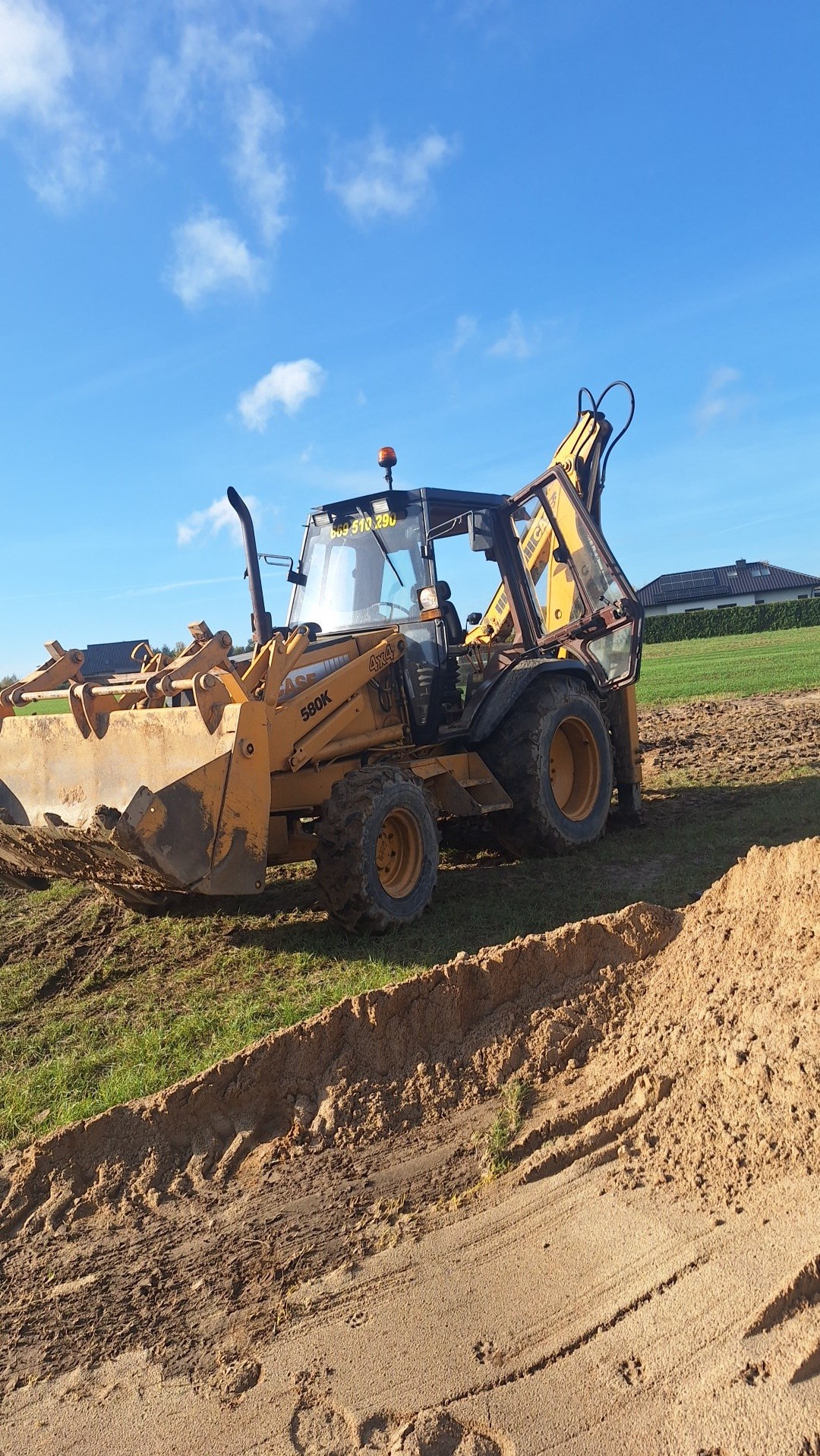 Żółta koparko-ładowarka Case 580K na trawiastym terenie z wykopanym rowem. Widoczne ślady opon i świeżo wykopana ziemia. Błękitne niebo w tle.