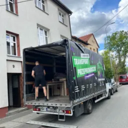 Czarno-zielona plandeka ciężarówki z napisem 'Transport Przeprowadzki' podczas załadunku mebli na ulicy przed budynkiem mieszkalnym.
