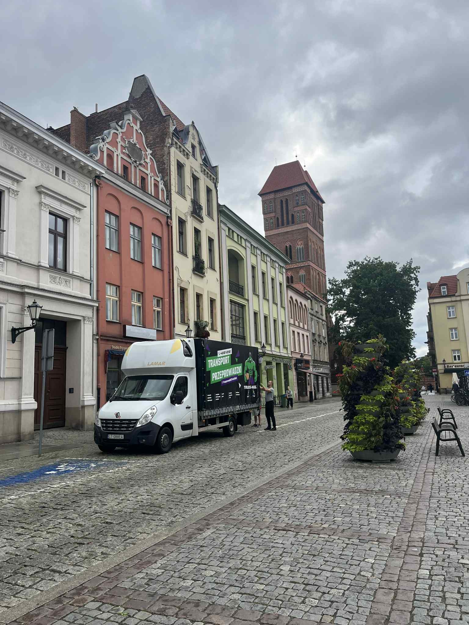 Biały bus z reklamą firmy przeprowadzkowej na tle kolorowych kamienic i wieży kościoła w Toruniu. Usługa transportowa w historycznym otoczeniu.