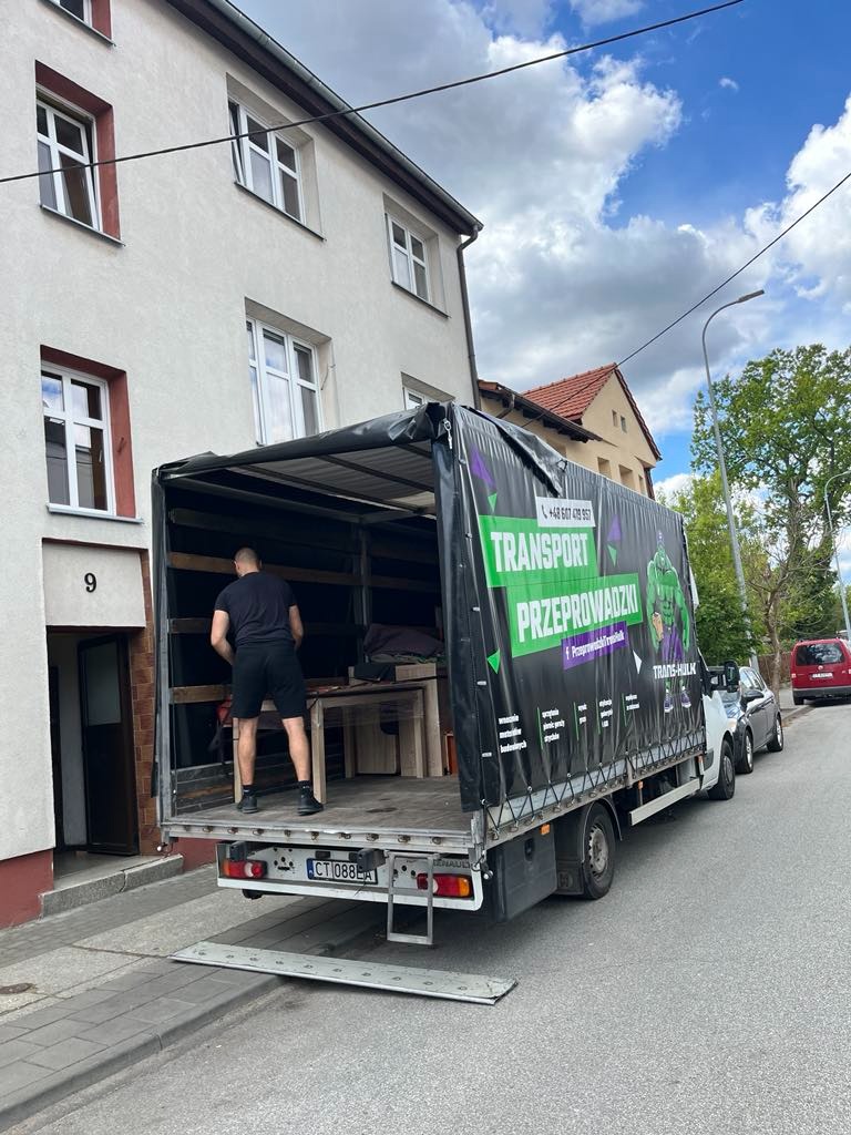Czarno-zielona plandeka ciężarówki z napisem 'Transport Przeprowadzki' podczas załadunku mebli na ulicy przed budynkiem mieszkalnym.