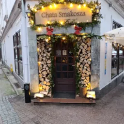 Wejście do restauracji 'Chata Smakosza' ozdobione drewnem, girlandami i światełkami, z czerwonymi kokardami. Uwagę przyciąga rustykalny wystrój i ciepłe oświetlenie.