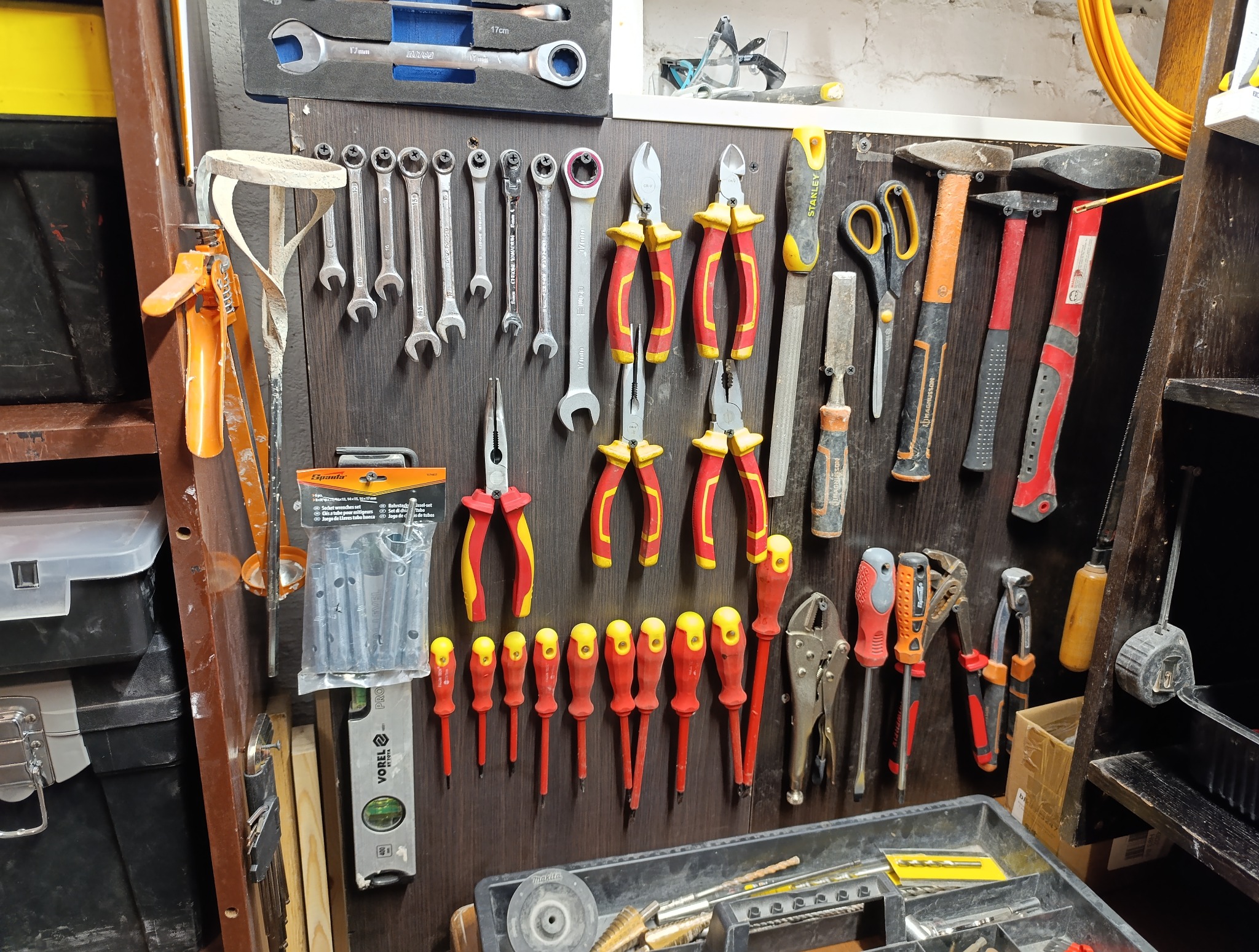 Ściana z narzędziami: klucze, kombinerki, wkrętaki (czerwono-żółte), młotki, poziomica Vorel, miara zwijana. Część narzędzi w skrzynce, inne wiszą na ścianie.