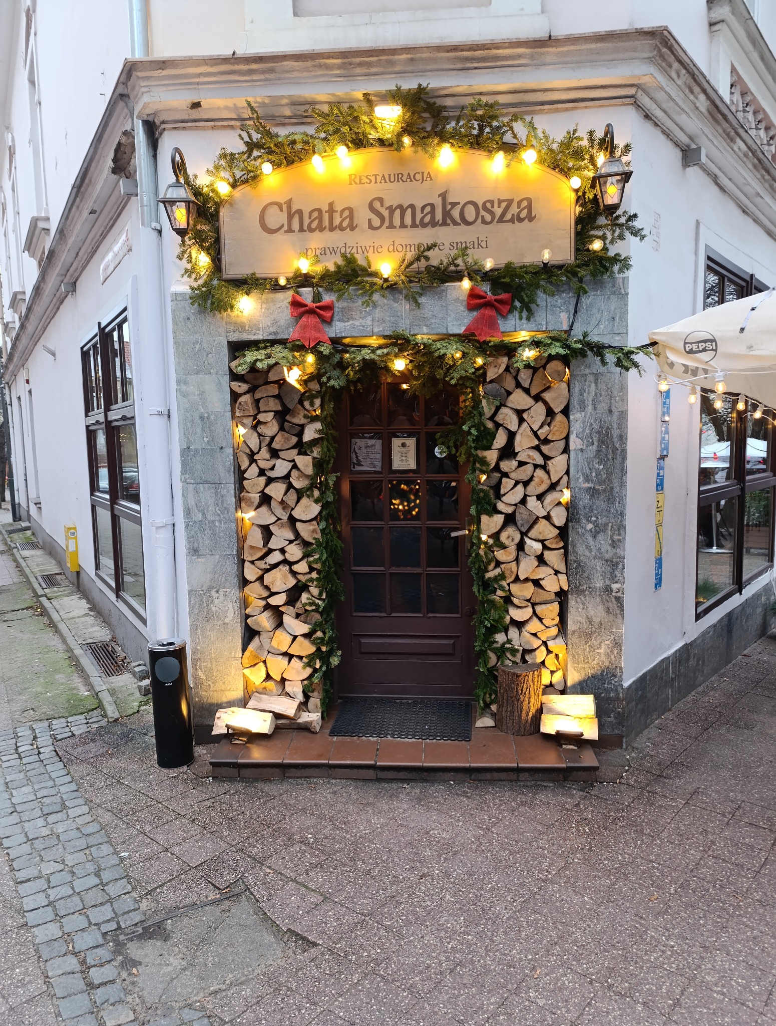 Wejście do restauracji 'Chata Smakosza' ozdobione drewnem, girlandami i światełkami, z czerwonymi kokardami. Uwagę przyciąga rustykalny wystrój i ciepłe oświetlenie.
