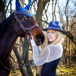 Ewa Bykowska Okiem Bykowskiej - Portret kobiety w niebieskim kapeluszu, trzymającej za uzdę brązowego konia w lesie. Uwagę zwraca dopasowany kolorystycznie strój konia i kobiety.