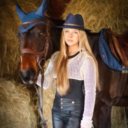 Ewa Bykowska Okiem Bykowskiej - Portret młodej kobiety w kapeluszu, stojącej obok konia z niebieskim czaprakiem na tle stogu siana. Ujęcie w stajni, naturalne światło.