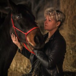 Ewa Bykowska Okiem Bykowskiej - Kobieta w czarnej kurtce, z krótkimi blond włosami, czule dotyka pyska ciemnego konia na tle stogu siana. Ujęcie w stajni, ciepłe światło.
