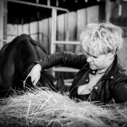 Ewa Bykowska Okiem Bykowskiej - Czarno-białe zdjęcie kobiety w skórzanej kurtce, głaszczącej konia w stajni. W tle drewniana konstrukcja i siano. Ujęcie z bliska, skupione na interakcji.