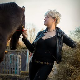 Ewa Bykowska Okiem Bykowskiej - Kobieta w czarnej kurtce i obcisłych spodniach karmi konia sianem na tle drewnianego płotu i nieba. Ujęcie z profilu, skupione na interakcji.