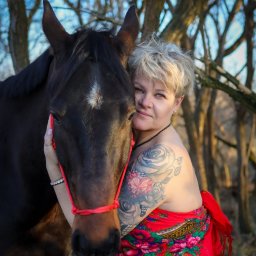 Ewa Bykowska Okiem Bykowskiej - Kobieta z tatuażami, w czerwonej chuście, obejmuje brązowego konia z białą gwiazdą na czole na tle drzew w naturalnym świetle.