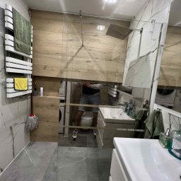 KAFELEK Adam Jaracz - Wykończona łazienka z przeszkloną kabiną prysznicową, grzejnikiem drabinkowym z ręcznikami, marmurowymi płytkami i drewnianymi panelami ściennymi.