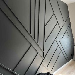 KAFELEK Adam Jaracz - Grafitowa ściana z geometrycznym wzorem z listew, subtelne oświetlenie, widoczny fragment podłogi i grzejnika. Dekoracja wnętrza.