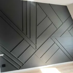 Nowoczesna ściana z geometrycznym wzorem z listew w odcieniach szarości, z widocznym fragmentem podłogi i gniazdkiem elektrycznym.