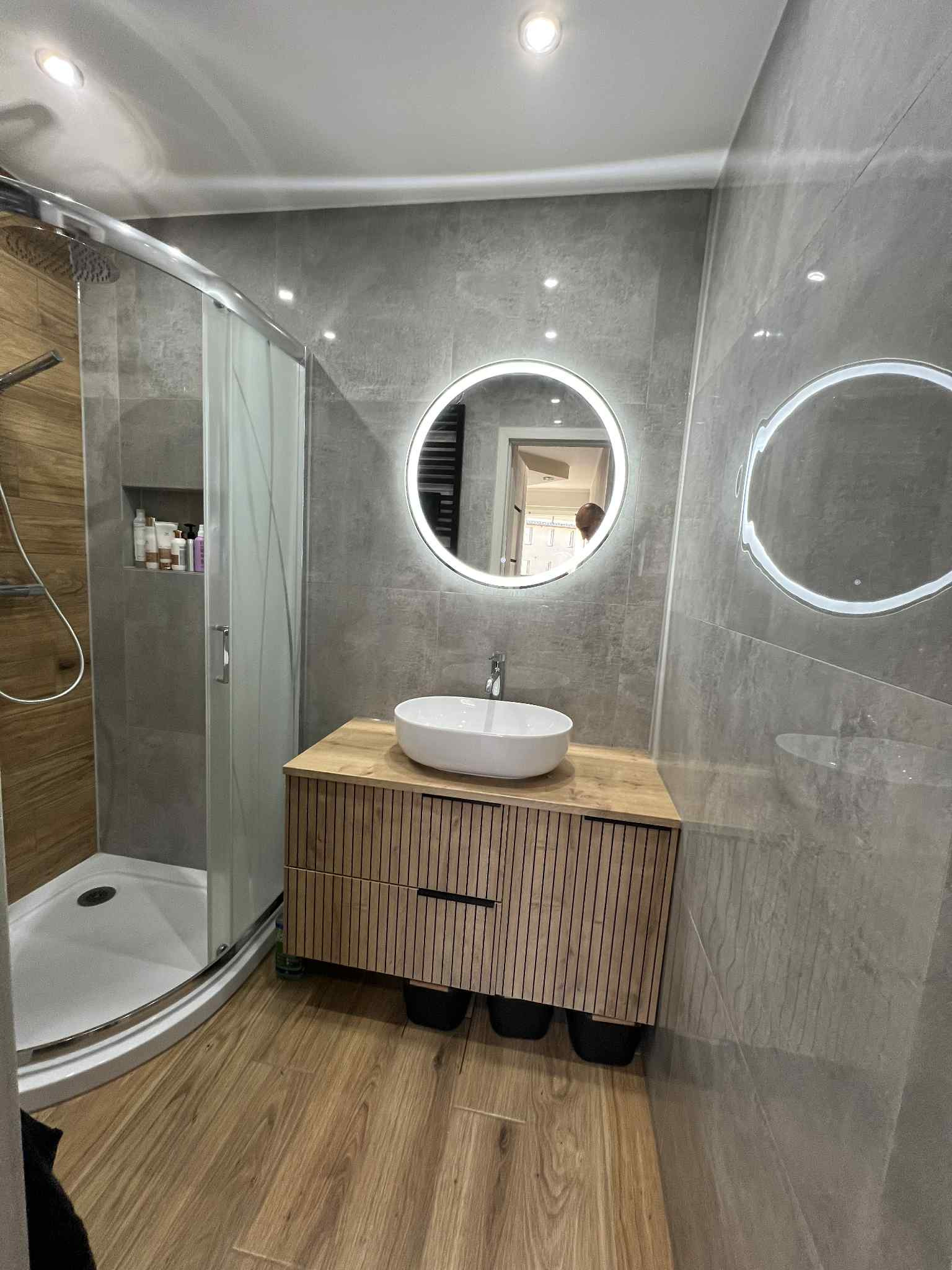 Nowoczesna łazienka po remoncie: kabina prysznicowa z drewnianymi panelami, umywalka nablatowa na szafce z lamelami, okrągłe lustra z podświetleniem LED, szare płytki.
