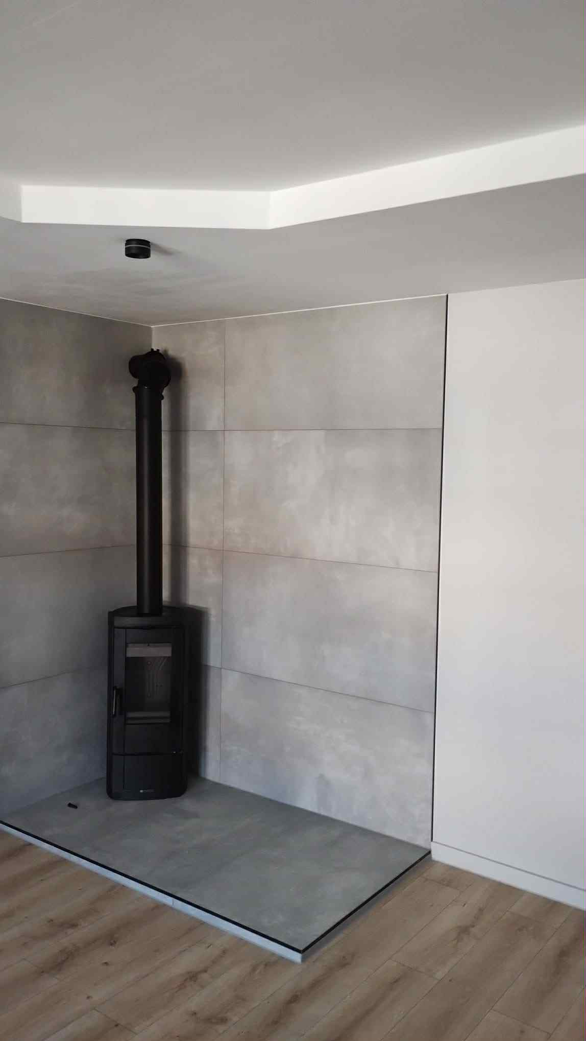 Nowoczesny piec wolnostojący z czarnym kominem na tle szarych, wielkoformatowych płytek. Minimalistyczne wykończenie wnętrza z drewnianą podłogą.