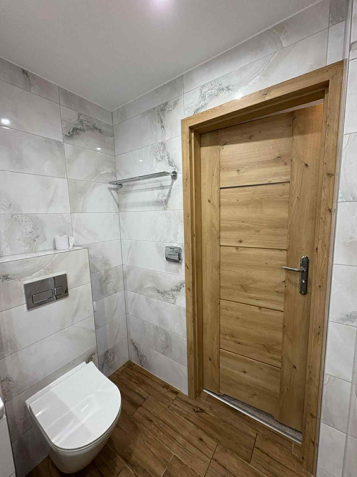 Nowoczesna łazienka z marmurowymi płytkami, drewnianą podłogą i drzwiami. Wisząca toaleta i chromowany wieszak na ręczniki dodają elegancji.