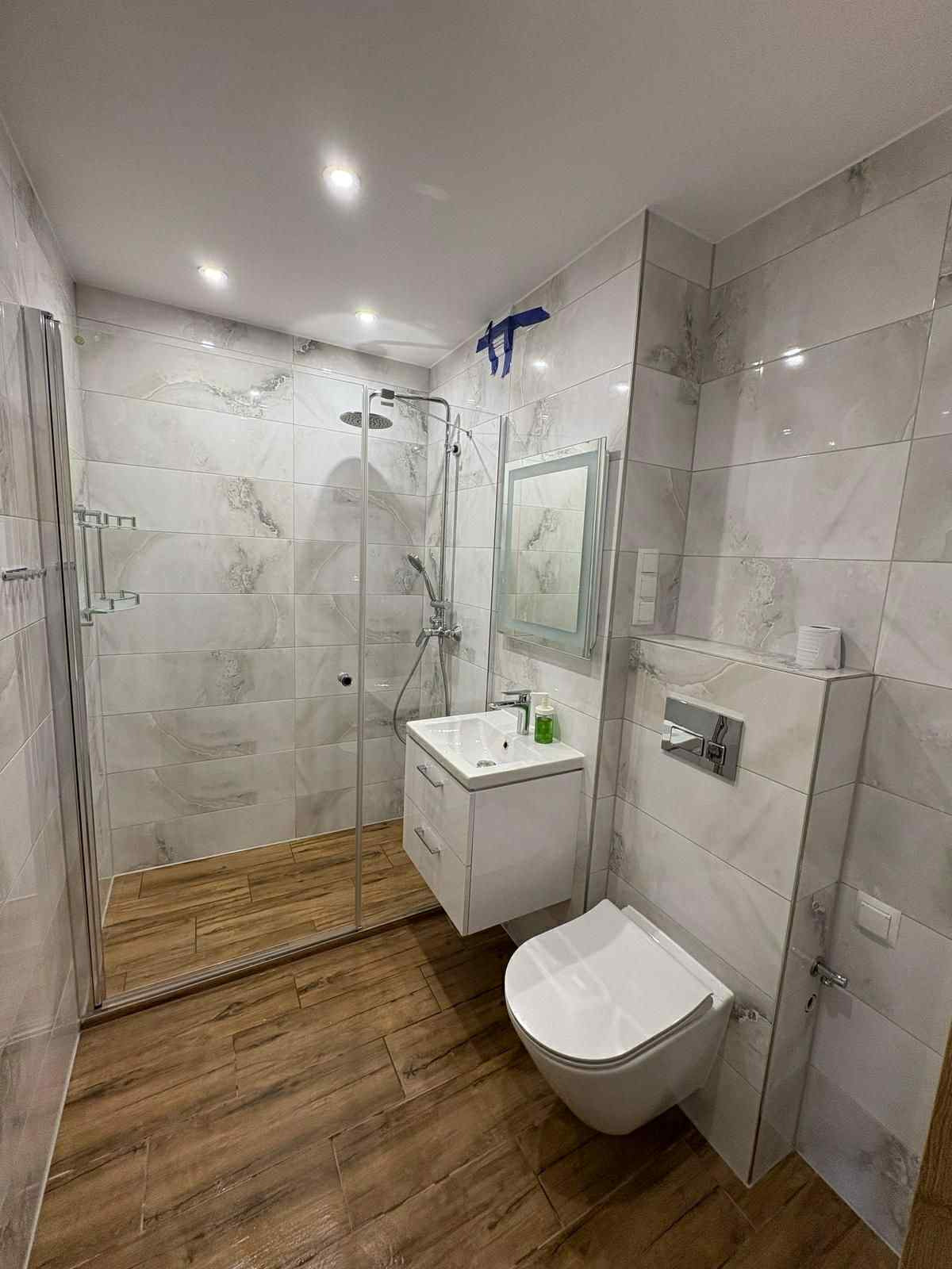 Wykończona łazienka z kabiną prysznicową, umywalką z szafką i podwieszaną toaletą. Na ścianach i podłodze płytki imitujące marmur i drewno. Niebieska taśma malarska na suficie.