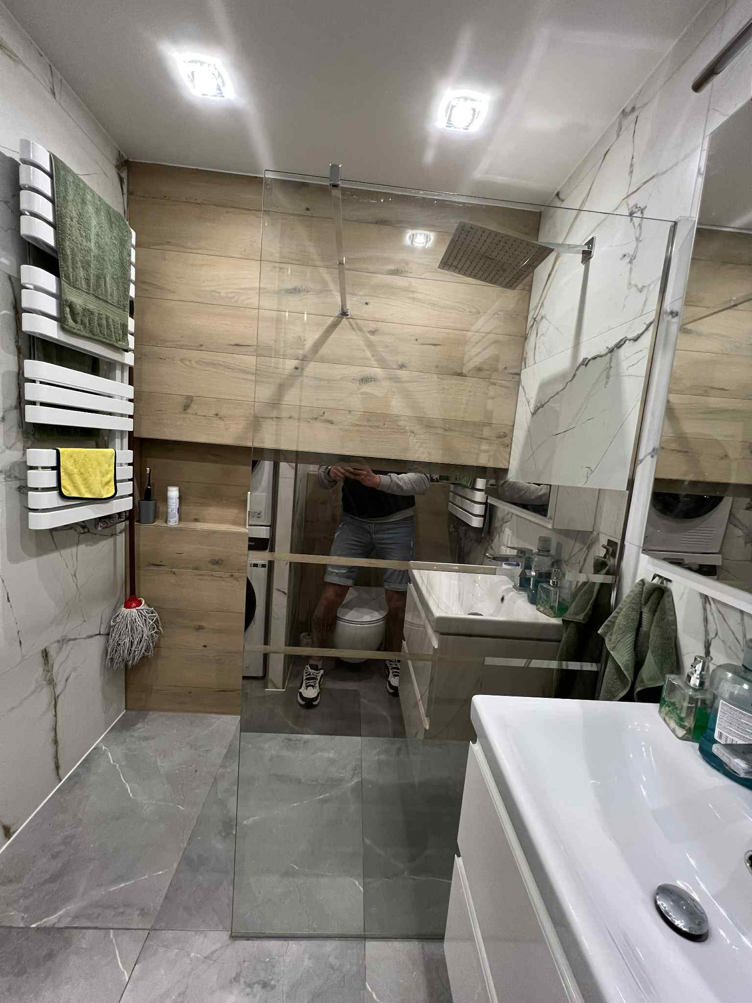 Nowoczesna łazienka z kabiną walk-in, marmurowymi płytkami i drewnianymi akcentami. Widoczny grzejnik drabinkowy z ręcznikami i szary mop. W lustrze odbija się osoba robiąca zdjęcie.