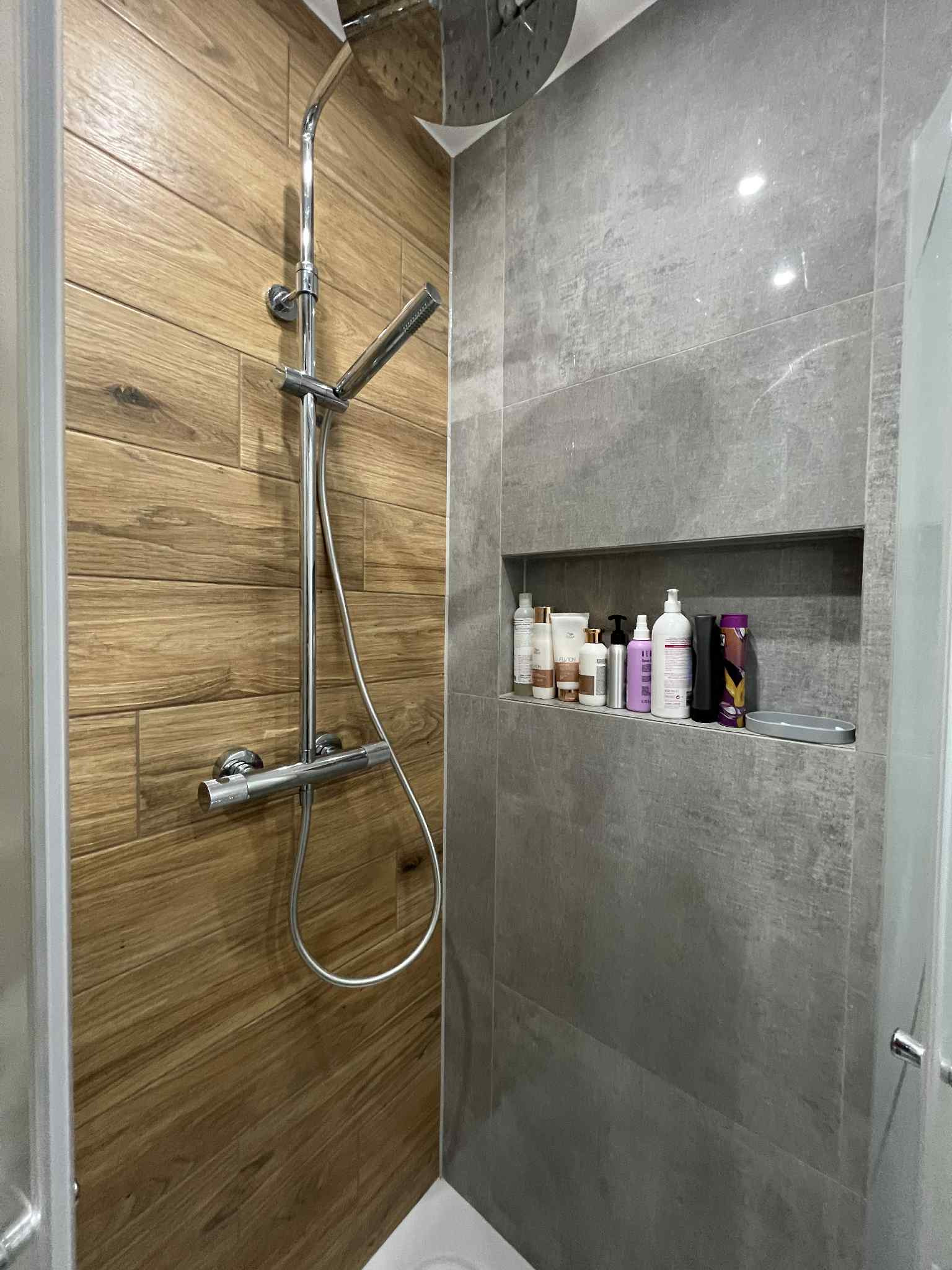 Nowoczesna kabina prysznicowa z drewnopodobnymi i szarymi płytkami, wnęka z kosmetykami. Chromowana armatura i deszczownica. Minimalistyczny design.