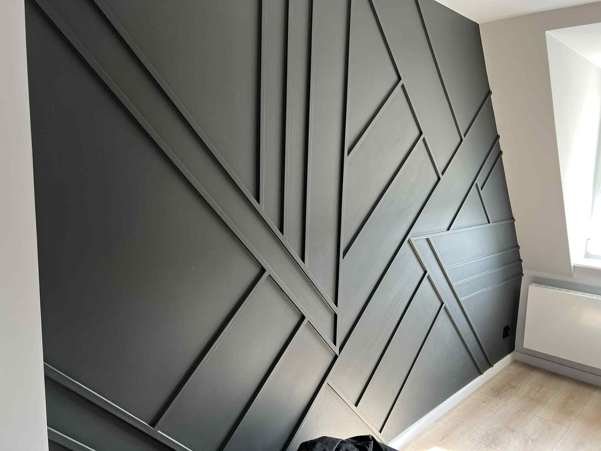 Grafitowa ściana z geometrycznym wzorem z listew, subtelne oświetlenie, widoczny fragment podłogi i grzejnika. Dekoracja wnętrza.