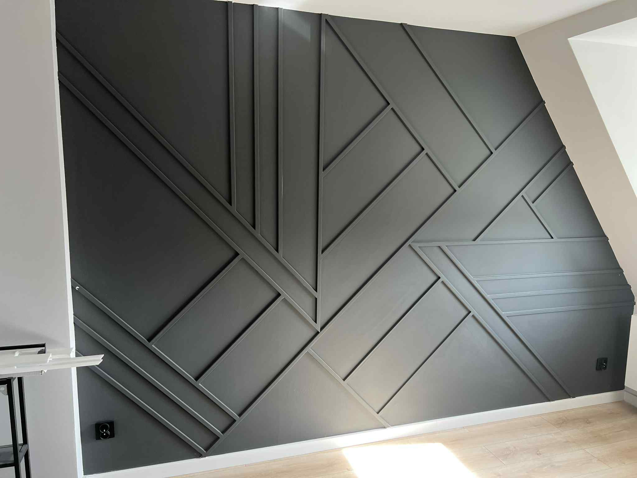Nowoczesna ściana z geometrycznym wzorem z listew w odcieniach szarości, z widocznym fragmentem podłogi i gniazdkiem elektrycznym.