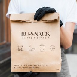 _bootelka Studio Graficzne - Papierowa torba z logo RU-SNACK Bistro Piekarnia, trzymana przez osobę w czarnych rękawiczkach. Widoczne grafiki: kawa, rogalik, napój, babeczka.