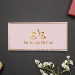 Elegancki, różowy voucher podarunkowy 'Harmonia Piękna' ze złotym logo i obramowaniem, leżący na ciemnym tle z dekoracją z białych kwiatów.