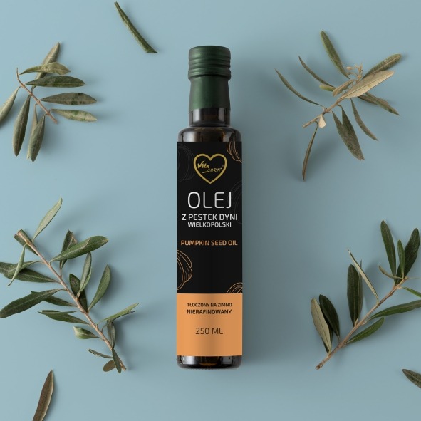 Butelka oleju z pestek dyni z etykietą Vita Zook na błękitnym tle, otoczona gałązkami oliwnymi. Minimalistyczny design, naturalne światło, widoczny detal etykiety.