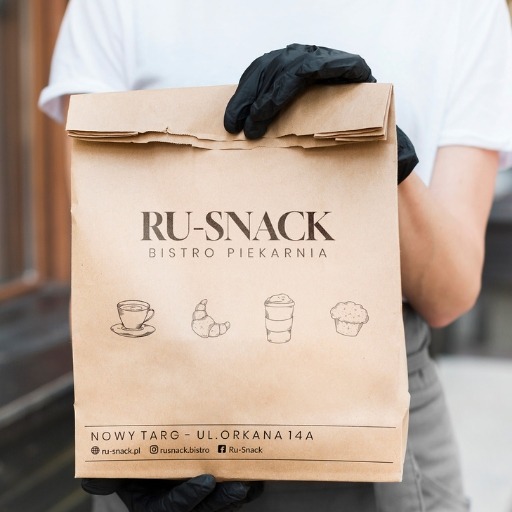Papierowa torba z logo RU-SNACK Bistro Piekarnia, trzymana przez osobę w czarnych rękawiczkach. Widoczne grafiki: kawa, rogalik, napój, babeczka.