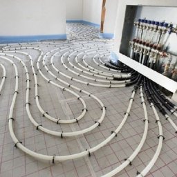 SK TECHNIK - Białe rury ogrzewania podłogowego ułożone spiralnie na siatce, z widoczną rozdzielnią w ścianie. Detal instalacji przed wylaniem posadzki.