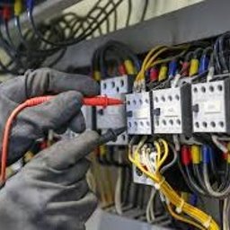 SK TECHNIK - Instalator w rękawicach diagnozuje obwód elektryczny za pomocą miernika, na tle rozdzielnicy z przewodami w kolorach: żółtym, niebieskim, czerwonym.