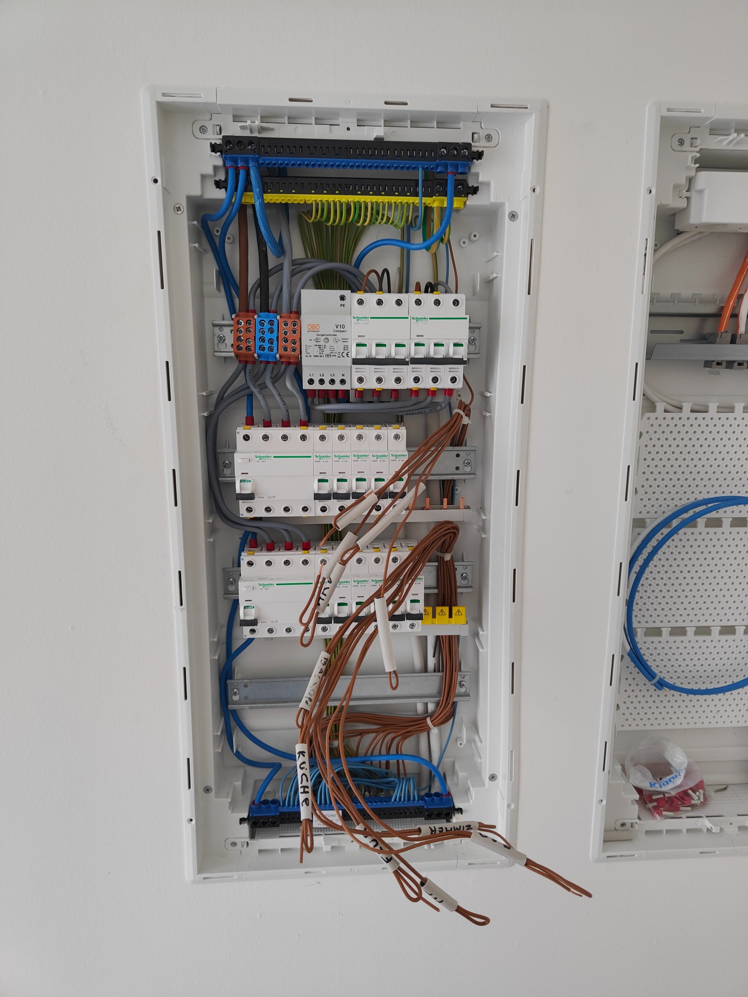 Rozdzielnica elektryczna w białej obudowie, z widocznymi przewodami oznaczonymi etykietami 'KUCHE' i 'ZIMMER', oraz zabezpieczeniami Schneider Electric.