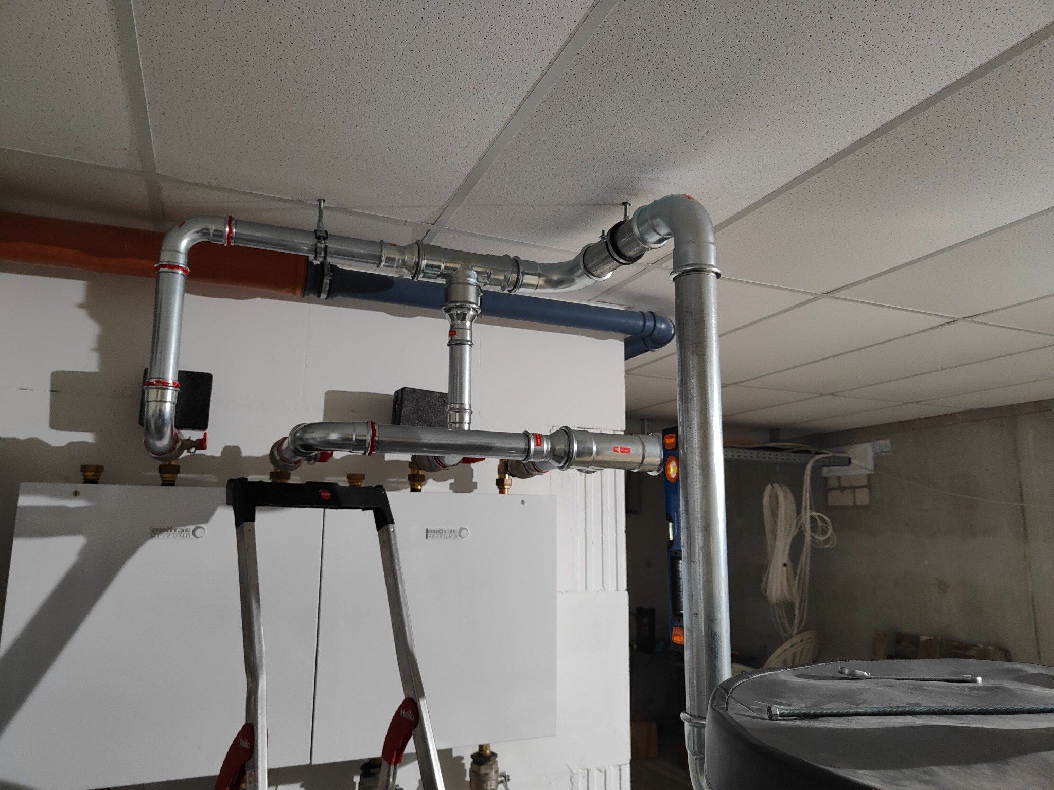 Instalacja grzewcza z dwoma piecami i rurami z czerwonymi złączkami, drabina oparta o piec, widoczne narzędzia i zbiornik. Biały sufit kasetonowy.