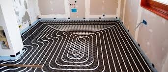 Białe rury ogrzewania podłogowego ułożone spiralnie na czarnej macie w surowym wnętrzu z widocznymi przewodami elektrycznymi i fragmentem okna.