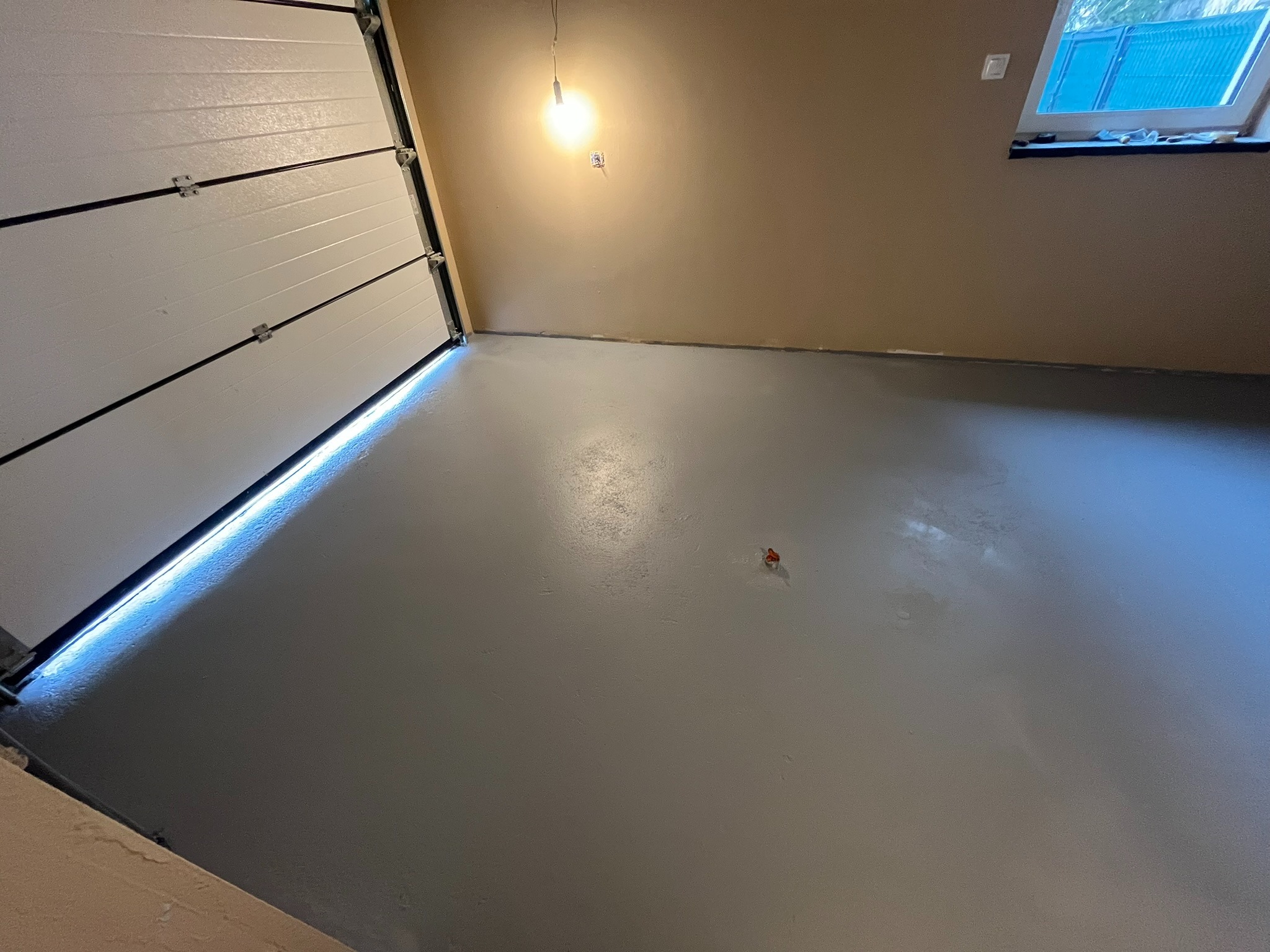 Szara, gładka posadzka żywiczna w garażu z otwartą białą bramą segmentową i oknem dachowym. Widoczna faktura i połysk posadzki.