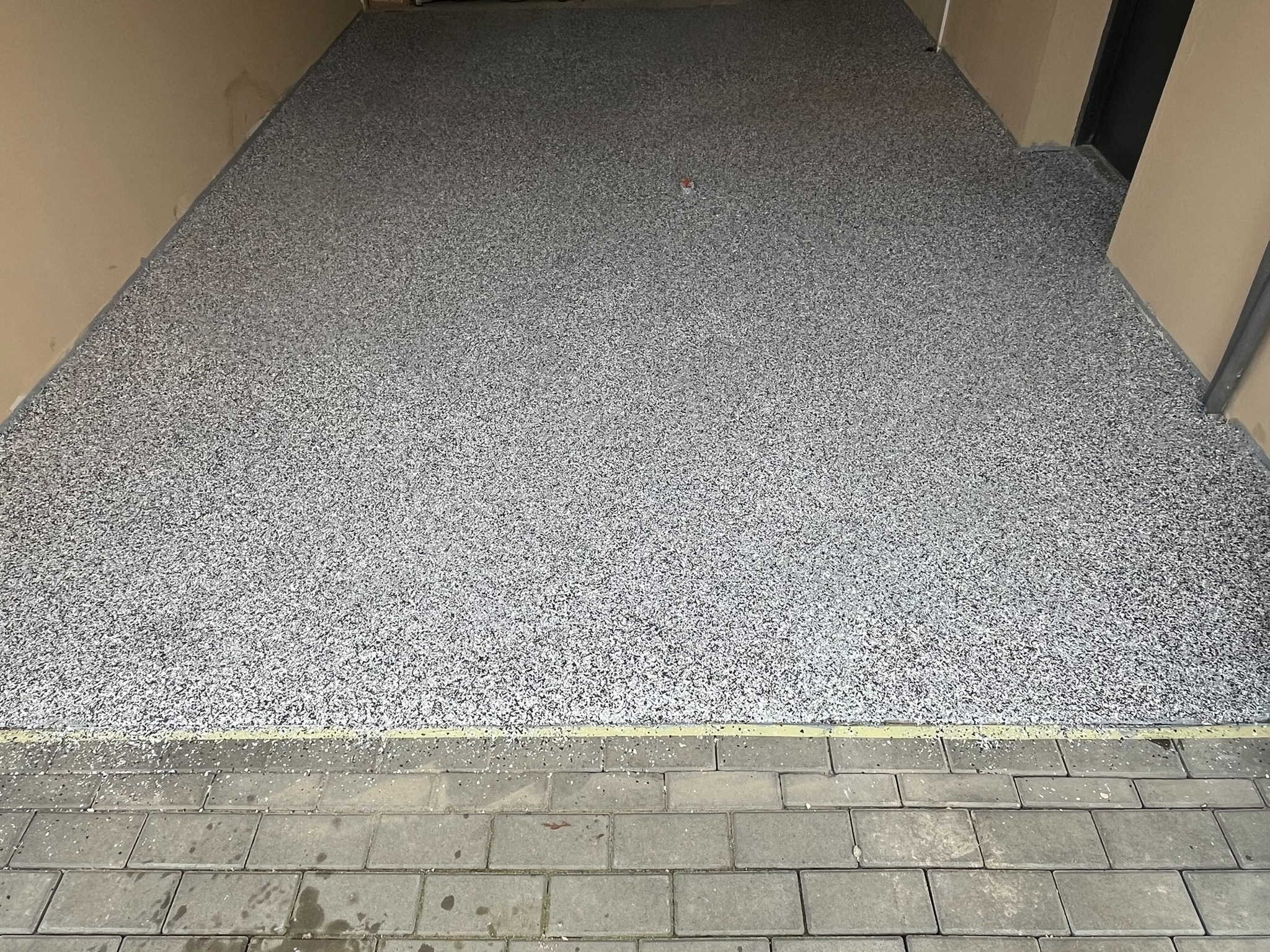 Szara posadzka żywiczna z drobnym wzorem w garażu, widoczna z góry, z fragmentem kostki brukowej przy wejściu. Ściany w odcieniach beżu.