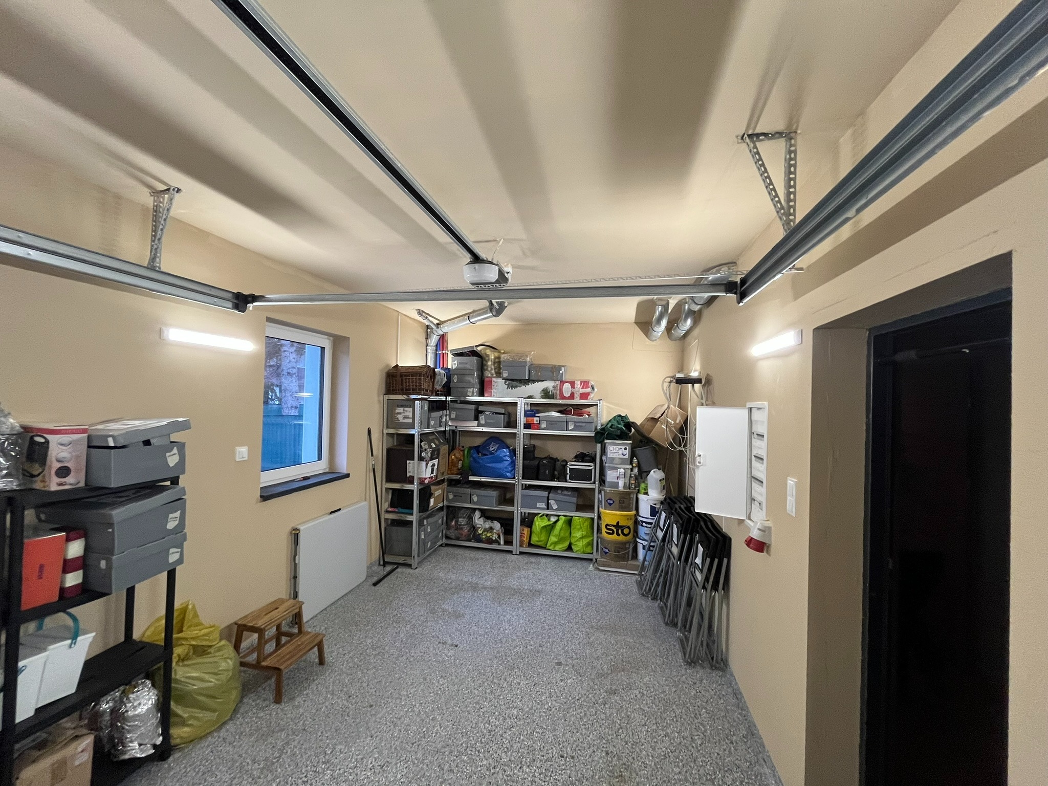 Garaż z posadzką żywiczną w szarym odcieniu. Widoczne regały z różnymi przedmiotami, okno z widokiem na zewnątrz i oświetlenie sufitowe.