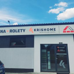 Krishome oddział w Opolu - Fasada budynku z szyldem 'Bramy, Okna, Rolety - Krisshome Salon Stolarki Krispol Dach Klinkier'. Niebieski samochód zaparkowany przed budynkiem. Błękitne niebo z chmurami w tle.