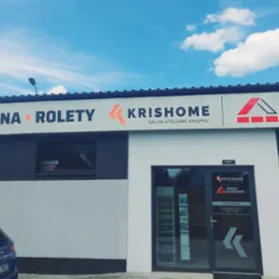 Fasada budynku z szyldem 'Bramy, Okna, Rolety - Krisshome Salon Stolarki Krispol Dach Klinkier'. Niebieski samochód zaparkowany przed budynkiem. Błękitne niebo z chmurami w tle.